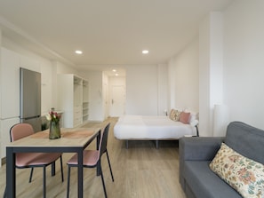 A compact room with a bed, a sofa, a dining table with chairs, a kitchen area with a refrigerator, and a wardrobe.