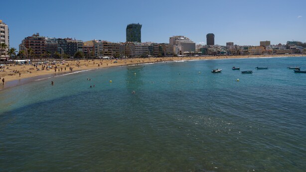 A coastal cityscape with a beach, clear blue water, and several boats.