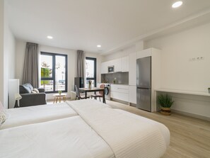 A modern hotel room with a large bed, a dining area, a kitchenette, and a view of the outdoors.