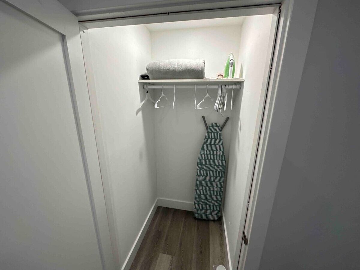 A small, narrow closet with a wooden floor, a hanging rack with clothes, and a shelf with a towel and an iron.