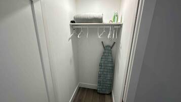 A small, narrow closet with a wooden floor, a hanging rack with clothes, and a shelf with a towel and an iron.