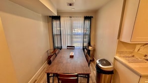 Un comedor con una mesa de madera, sillas, un cubo de basura y una ventana con cortinas.