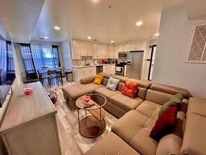 A modern living room with a sectional sofa, a coffee table, and a kitchen in the background. - The Brand New Suite: 2BR with Parking (Paterson)