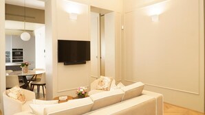 Een moderne woonkamer met een zithoek, een flatscreen televisie aan de muur en een eethoek zichtbaar door een open deur.