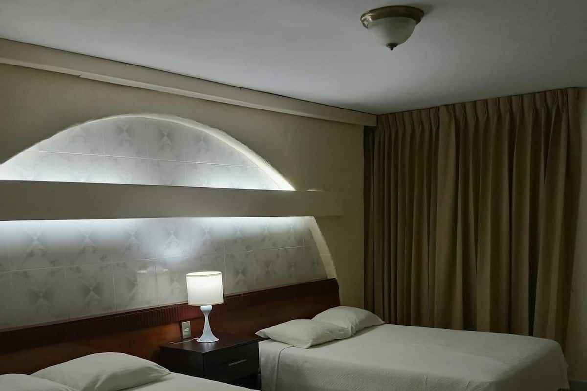 A hotel room with two beds, a nightstand, a lamp, a ceiling light, and a large window with curtains.