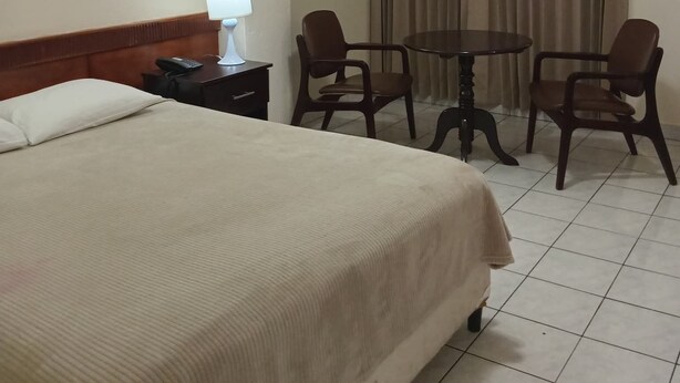 A hotel room with a bed, a bedside table with a lamp, a telephone, two chairs, a small round table, and a curtain. - Hotel Estrella (Managua)