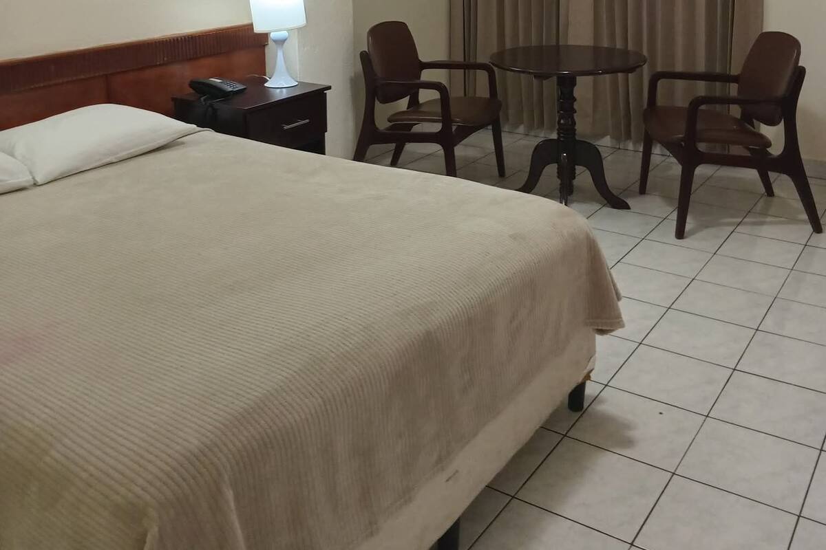 A hotel room with a bed, a bedside table with a lamp, a telephone, two chairs, a small round table, and a curtain.