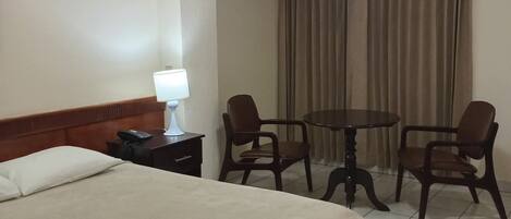A hotel room with a bed, a bedside table with a lamp, a telephone, two chairs, a small round table, and a curtain.
