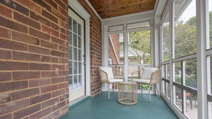 A covered porch with brick walls, white-framed windows, wicker chairs, and a small table. - Charming 2-bedroom house with WiFi, AC in brilliant Geneva (Geneva)