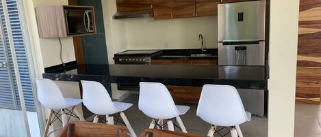 Una cocina moderna con armarios de madera, un refrigerador de acero inoxidable y un horno empotrado. Hay cuatro taburetes blancos y una mesa de comedor de madera.