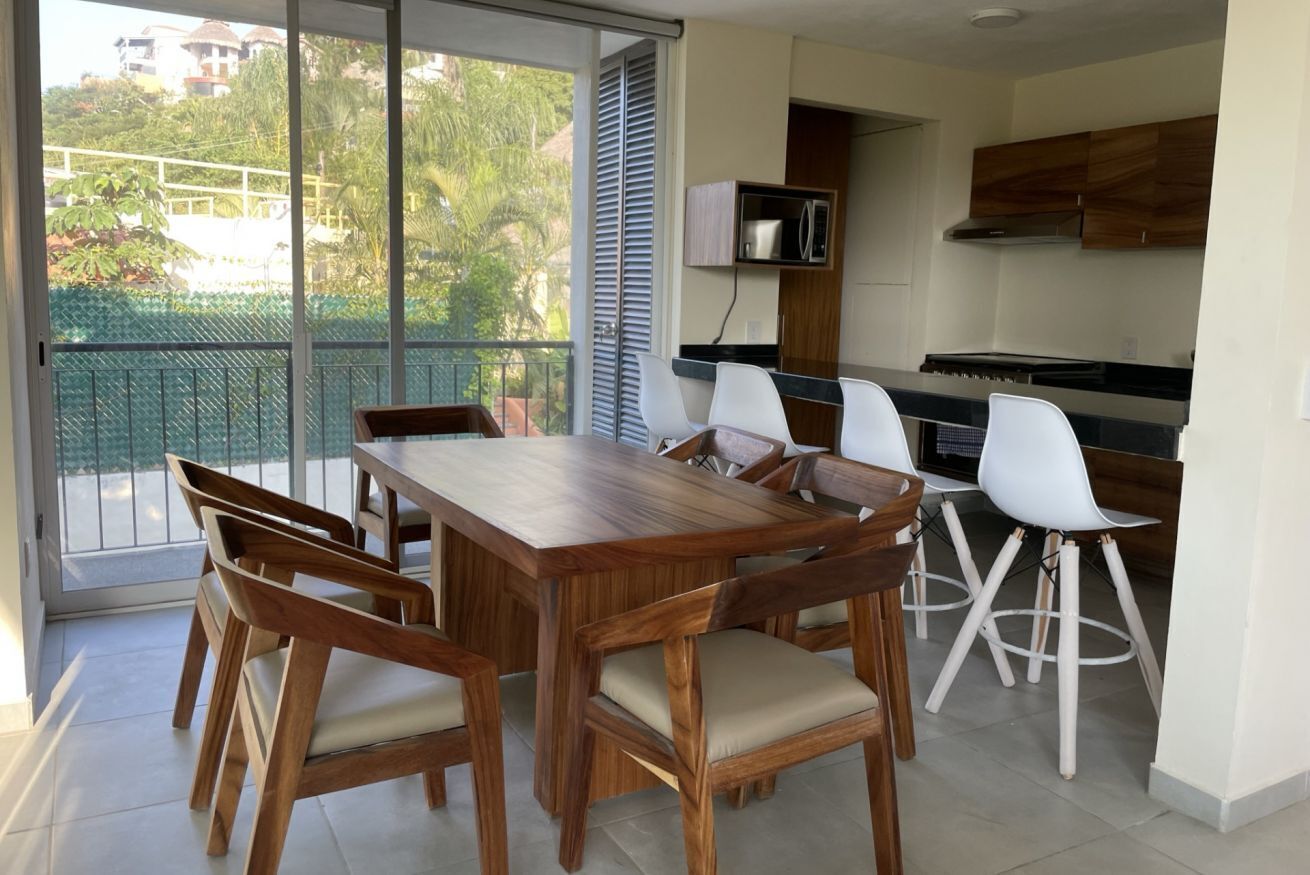 A modern kitchen with a dining area, large windows, and a view of greenery.