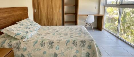 A bedroom with a bed, a wooden wardrobe, a desk, and a large window with a view of greenery.