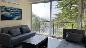 Una sala de estar moderna con un sofá gris, una mesa de centro negra y un ventanal con vistas a follaje y un paisaje distante.