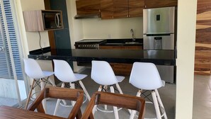 Una cocina moderna con armarios de madera, un refrigerador de acero inoxidable y un horno empotrado. Hay cuatro taburetes blancos y una mesa de comedor de madera.