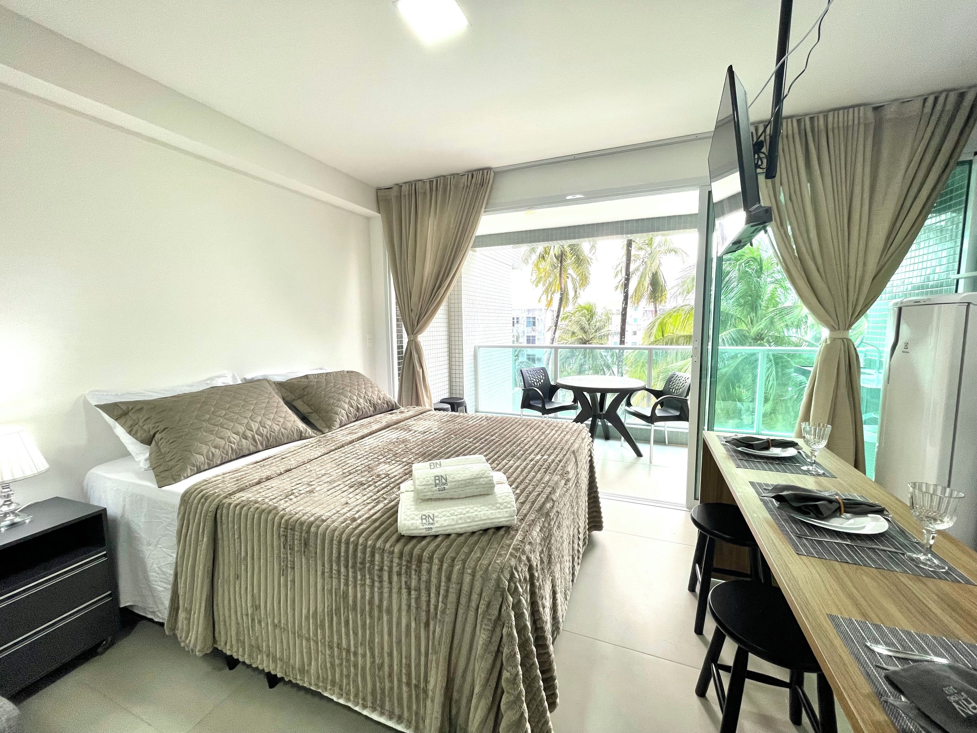 A hotel room with a bed, a desk with a chair, a TV, and a balcony with a table and chairs.