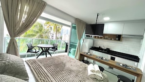 A modern bedroom with a large bed, a balcony with a view of palm trees, a small kitchen area, and a dining table. - RN Studio Premium 103 - Vista Mar (Maceió)