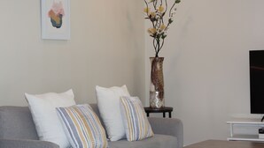 A modern living room with a grey sofa, two patterned cushions, a framed artwork on the wall, a tall vase with flowers, and a flat-screen TV on a white stand. - Cozy 3 Bedroom/Great Location (Arlington)