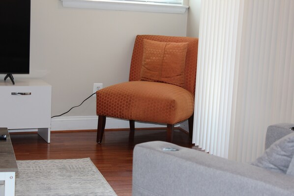 A living room with a television on a stand, a wooden floor, a grey sofa, and an orange armchair with a patterned cushion. - Cozy 3 Bedroom/Great Location (Arlington)