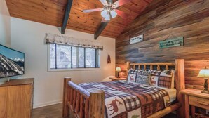 A cozy cabin-style bedroom with a wooden bed, a window with blinds, a ceiling fan, and a wall-mounted sign.