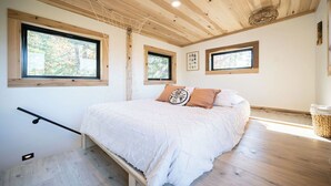 Een slaapkamer met een houten plafond, een groot bed met witte beddengoed, twee ramen en een klein wandplankje.