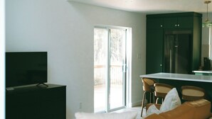 A modern living room with a sofa, a flat-screen TV, and a kitchen area with a refrigerator and bar stools. - Cozy Cabin Getaway (St. Ignace)