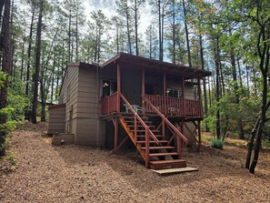 A cabin with a wooden deck and stairs leading to a balcony, surrounded by a forest.