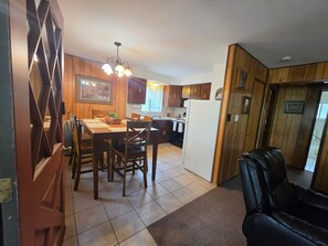 A kitchen with wooden cabinets, a dining table with chairs, a refrigerator, and a black leather sofa.