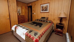 Quarto com paredes de madeira, uma cama com colcha temática de urso, uma mesa de cabeceira com abajur e um quadro na parede.