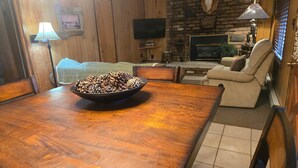 A wooden dining table with a bowl of pine cones, a television, a leather sofa, and a stone wall with a mounted animal skull. - Northwoods (#9) Cabin in the Pines (Pinetop-Lakeside)