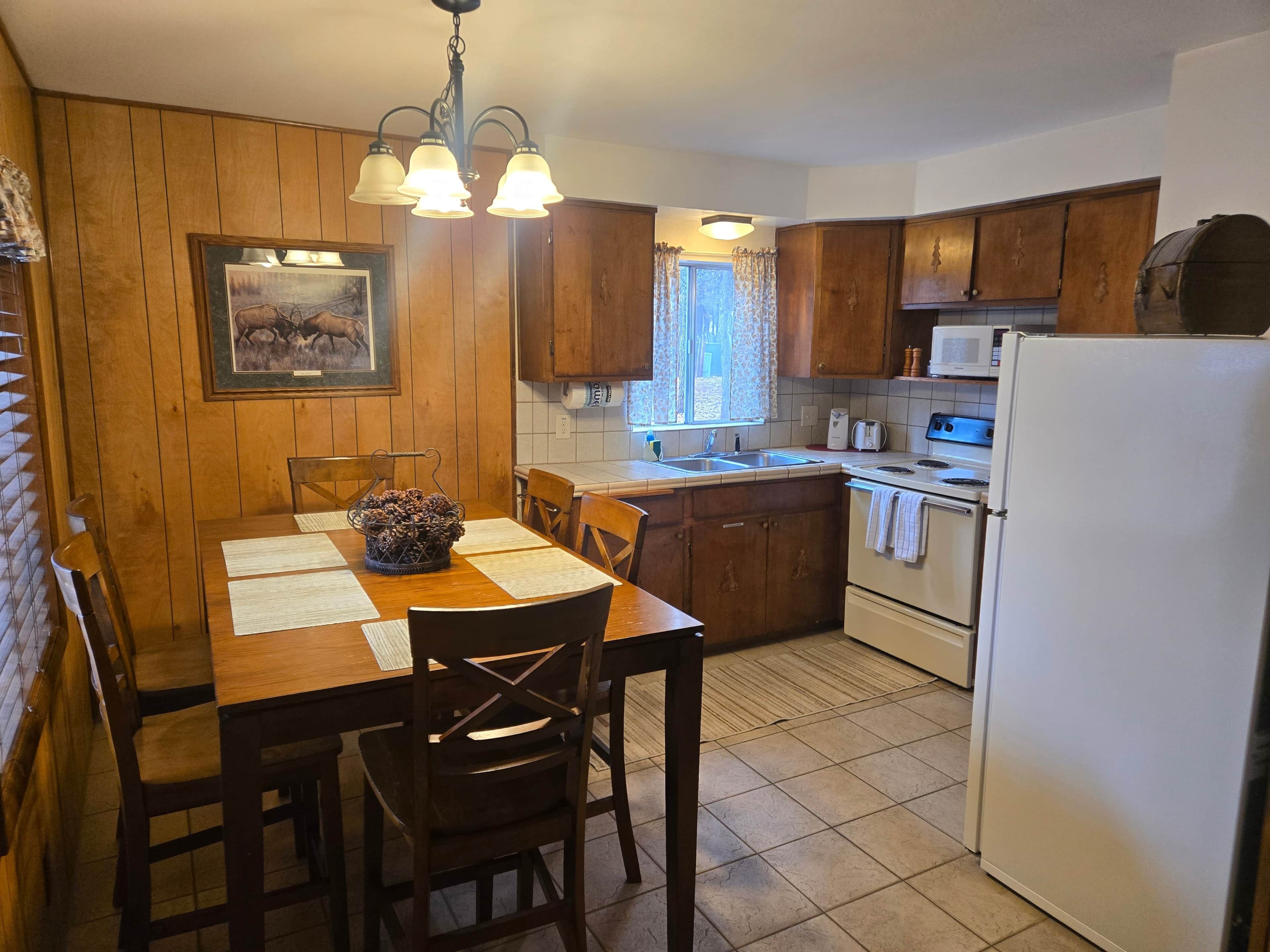 A kitchen with wooden cabinets, a dining table with chairs, a chandelier, a microwave, and a refrigerator.
