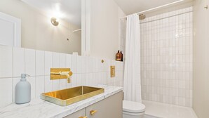 Ein modernes Badezimmer mit goldener Spüle, weißen Fliesen, Toilette und Duschbereich.