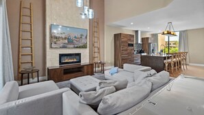 Ein modernes Wohnzimmer mit Kamin, grauen Sofas, einem Fernseher und einer Küche im Hintergrund.