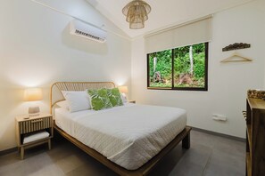 A bedroom with a bed, bedside table, lamp, air conditioning unit, window with a view of greenery, and a wall-mounted light fixture.
