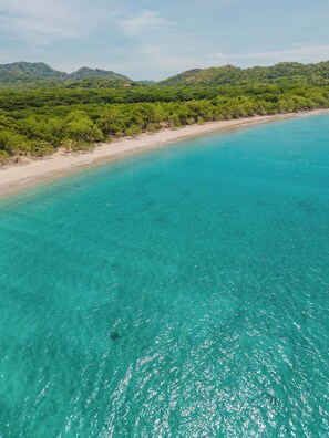 A coastal landscape with a clear turquoise sea, sandy beach, and dense green vegetation on the hills.