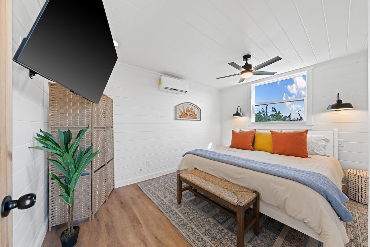 A bedroom with a bed, a ceiling fan, a window with a view, a wall-mounted TV, and a potted plant.