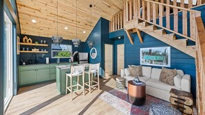 Un vaste espace de vie agrémenté d’un escalier en bois, une cuisine avec des meubles verts et un coin salon confortable avec un tapis à motifs.