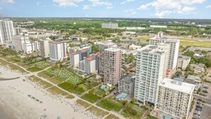 A high-rise residential building complex with multiple apartment buildings, a beach area with umbrellas, and a clear sky. - Oceanfront Lux at Palms Resort Views and Amenities (Myrtle Beach)