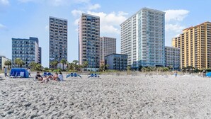 A beach with high-rise buildings in the background, including a hotel with multiple floors and balconies. - Oceanfront Lux at Palms Resort Views and Amenities (Myrtle Beach)