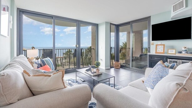 A spacious living area with a large sliding glass door leading to a balcony with a view of the ocean. There is a flat-screen TV mounted on the wall and a coffee table with a laptop and decorative items. - Oceanfront Lux at Palms Resort Views and Amenities (Myrtle Beach)