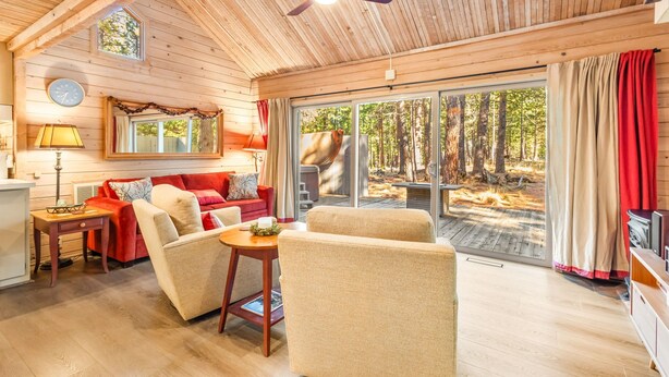 Interior acogedor de una cabaña con techo de madera, ventilador de techo, sofá rojo, sillones color beige, una mesita y un ventanal con vistas al bosque.