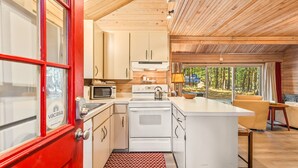 Cocina compacta con armarios blancos, microondas y estufa. Se observa una puerta roja con ventanal y vistas a un bosque a través de amplios ventanales.