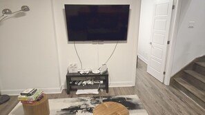 A living room with a flat-screen TV mounted on the wall, a small black entertainment unit with various electronic devices, a wooden side table with books, and a black and white rug.