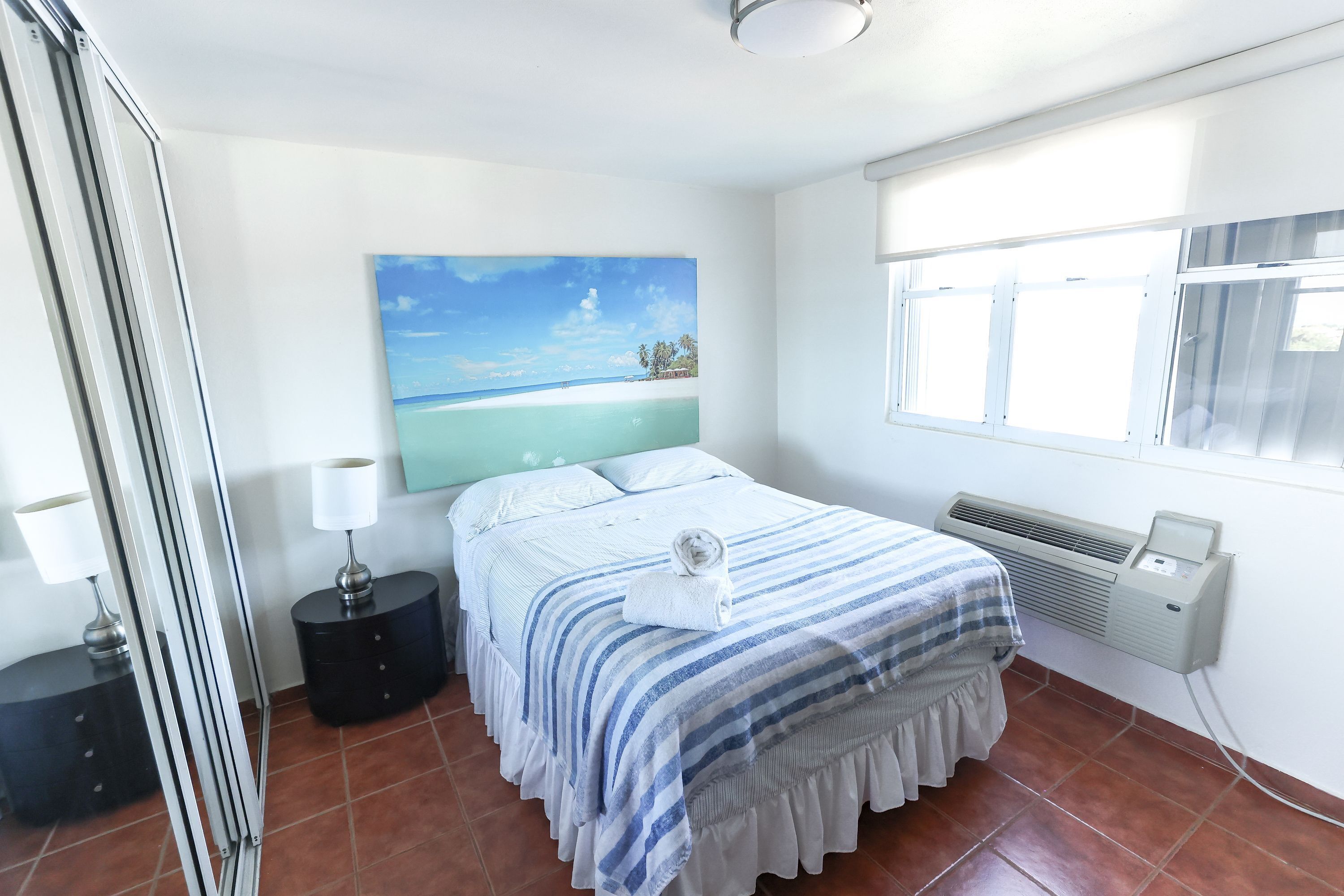 Een slaapkamer met een bed, nachtkastje, een wandairconditioner en een raam met jaloezieën.