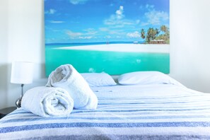 A neatly made bed with a striped blue and white bedspread, a rolled white towel, and a painting of a beach scene on the wall behind. - Relaxing Poolside Getaway With Balcony (Fajardo)