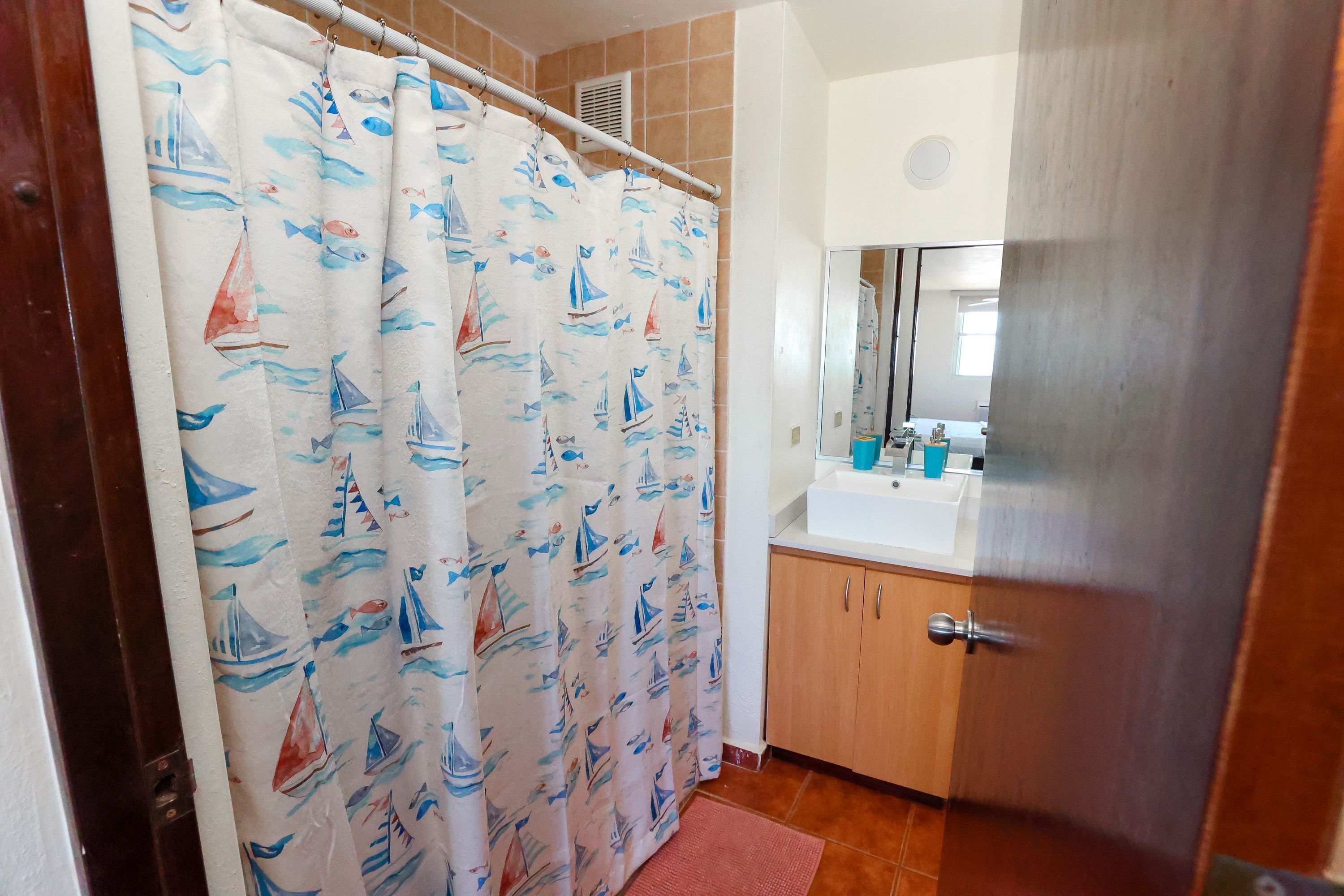 A bathroom with a shower curtain featuring sailboats and fish, a sink with toiletries, and a wooden cabinet.