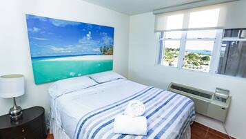 A hotel room with a bed, a nightstand, a lamp, a window with a view, and a painting on the wall.