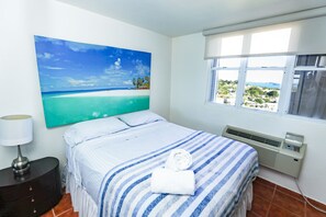 Habitación de hotel con cama, mesita de noche, lámpara, ventana con vistas y un cuadro en la pared.