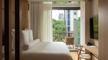 Habitación de hotel moderna con una cama grande, un escritorio, una silla y un balcón con vistas a edificios y zonas verdes.