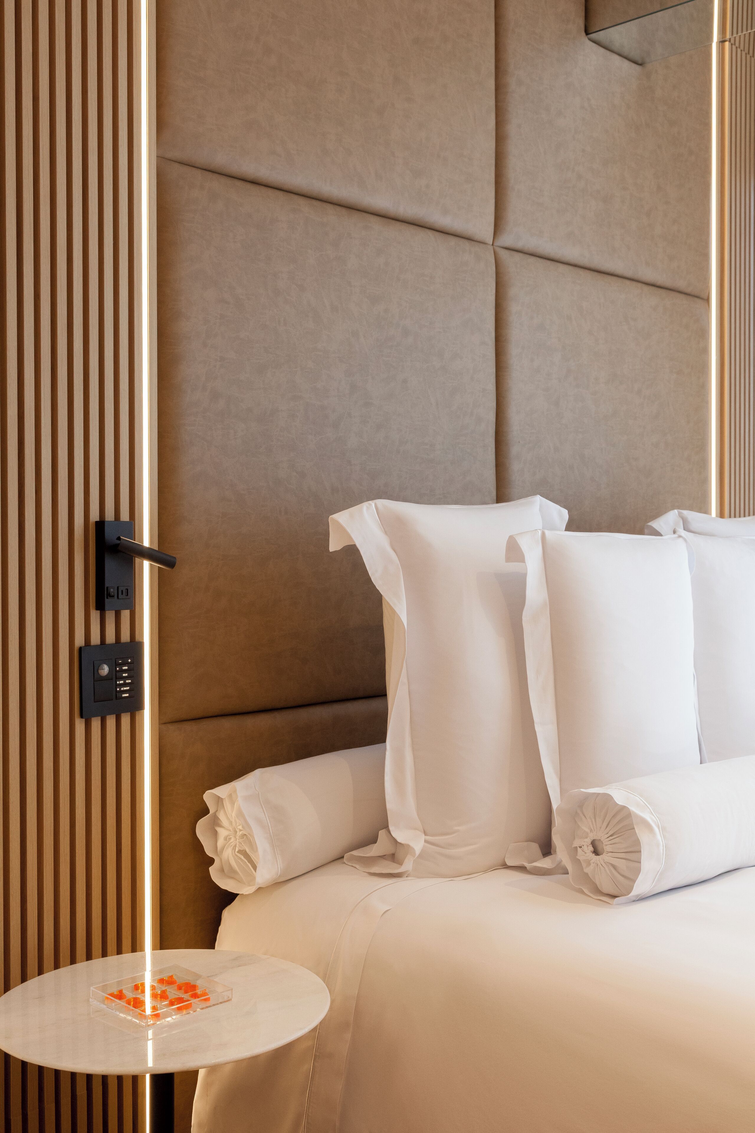 A hotel room with a modern design, featuring a large bed with white linens, a round side table with a clear glass object, and a wall-mounted control panel.