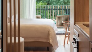 Ein Schlafzimmer mit einem Bett, einem Holzstuhl, einem kleinen Tisch und einem Balkon mit Blick auf Grünflächen.